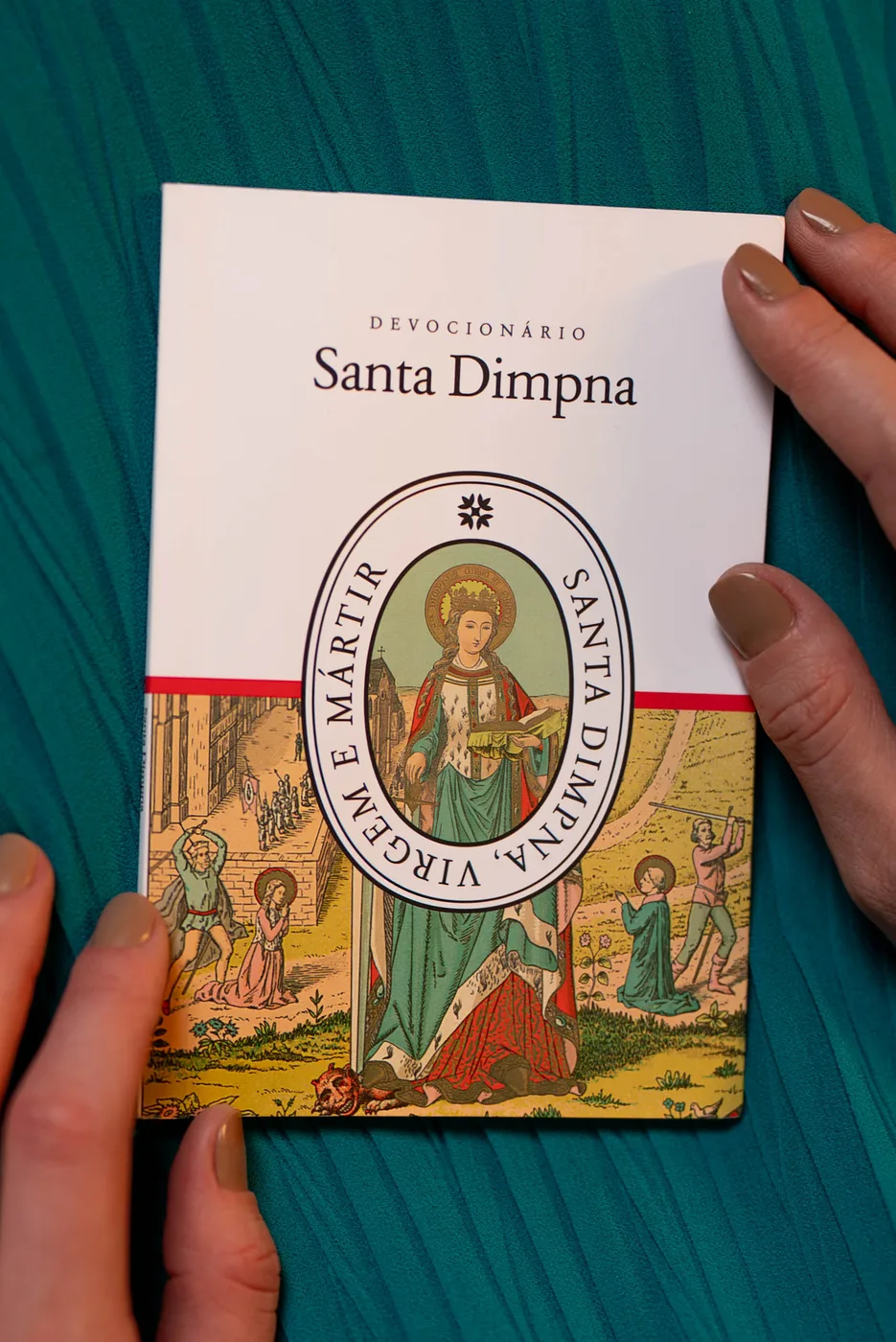 Mãos segurando o Devocionário a Santa Dimpna sobre tecido verde; capa branca com faixa vermelha e gravura da santa